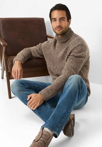 Een bruine, gebreide coltrui met een gestructureerd patroon, gecombineerd met blauwe jeans en lichtbruine suède schoenen. Zittend naast een bruine stoel.