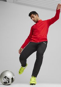 Rote Sportjacke, schwarze Hose und strahlend gelbe Fußballschuhe. Der Spieler ist in Bewegung, in der Nähe eines silbernen Fußballs mit einem schwarzen Logodetail.
