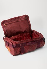 Sac de voyage en tissu rouge avec une fermeture éclair supérieure, une poche en maille intérieure et un marquage extérieur. Il possède des poignées robustes et des accents orange.