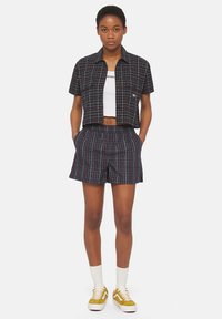 Camisa de cuadros negra recortada con dos bolsillos frontales, combinada con pantalones cortos de cuadros a juego. Top blanco debajo, zapatillas amarillas.