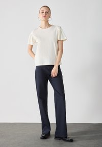 Top crema a maniche corte con dettagli arricciati, abbinato a pantaloni a gamba larga blu navy e scarpe lucide nere su una superficie grigia testurizzata.