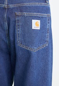 Denim jeans in dark blue with a pointed back pocket design. Features a Carhartt logo patch in white and orange near the pocket.