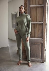 Maslinasto zelena pletiva pulover s visokim ovratnikom i uskim manžetama, uparen s odgovarajućim uskim hlačama od mekog materijala, obučen bosi na neutralnoj pozadini.