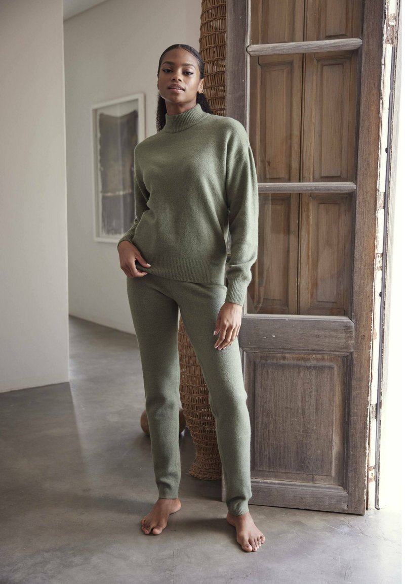 Maslinasto zelena pletiva pulover s visokim ovratnikom i uskim manžetama, uparen s odgovarajućim uskim hlačama od mekog materijala, obučen bosi na neutralnoj pozadini.