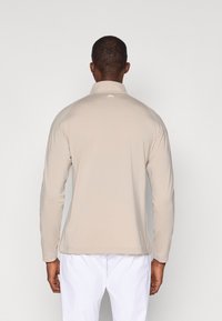 Koszula z długim rękawem w jasnobeżowym kolorze, z kołnierzykiem. Posiada minimalistyczny design i miękką fakturę, z małym logo poniżej szyi.