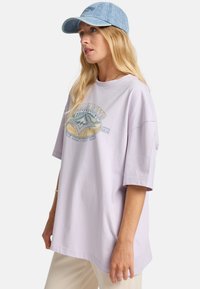 Camiseta oversized de color púrpura claro con un estampado gráfico que presenta olas y una marca. Material de algodón suave y corte relajado. Complementada con una gorra de denim.
