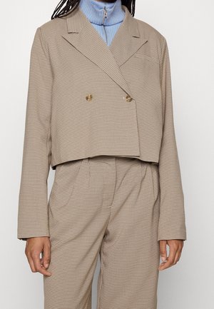 Person wearing beige checkered blazer and matching high-waisted trousers over a blue zip-up ribbed turtleneck sweater.