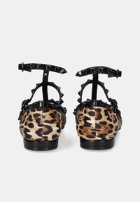 Par de zapatos planos con estampado de leopardo y correas negras con tachuelas en el tobillo, vistos desde la parte trasera sobre un fondo liso.