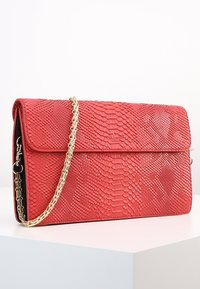 Červená kabelka z hadí kůže s překlopným víkem a zlatým řetízkovým popruhem, obsahující texturovaný povrch a diskrétně embosované logo na přední straně.