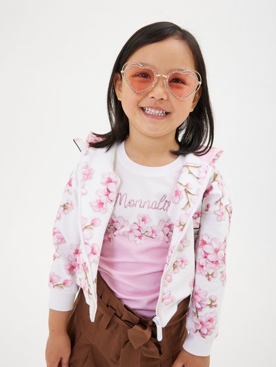 Sudadera con cremallera floral con flores rosas, superpuesta a una camiseta de color rosa claro con el texto "Mornala", combinada con gafas de sol rosas en forma de corazón y pantalones cortos marrones.