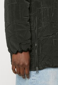 Svart texturerad jacka med blixtlåsstängning och elastiska ärmslut, samt en synlig sömnadsmönster. Modellens hand vilar på ett jeanstyg.