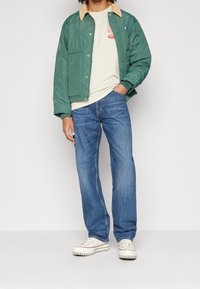 Zelená prošívaná bunda s béžovým límcem, nošená přes krémovou košili, v kombinaci s modrými džínami a béžovými teniskami, představující ležérní vzhled.