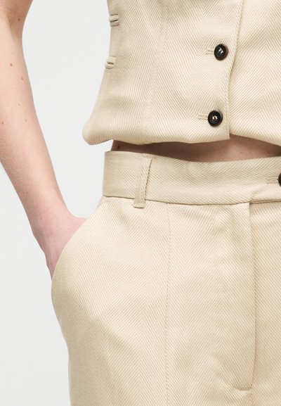 Άτομο που φοράει μπεζ υφασμάτινο παντελόνι και ταιριαστό γιλέκο με μαύρα κουμπιά, με το χέρι στην τσέπη, μπροστά σε μονόχρωμο φόντο.