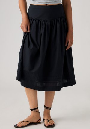 Femme portant une jupe noire longueur genou avec une taille froncée et des sandales plates noires à brides, vue du ventre aux pieds.
