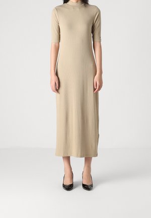 Femme portant une robe midi en maille côtelée beige avec manches longueur coude et des escarpins noirs à bout pointu sur fond blanc.