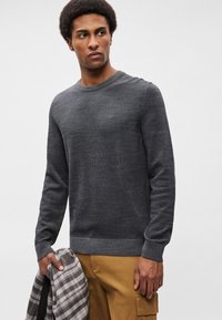 Ciemnoszara sweter z dzianiny z okrągłym dekoltem i ściągaczami na rękawach. Model trzyma w dłoniach koszulę w kratę w odcieniach szarości i bieli.