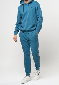 Tyrkysová mikina s kapucí a odpovídající tepláky z měkkého, pružného materiálu, s přední kapsou a vyšitým logem. Bílá sportovní obuv doplňuje celý outfit.