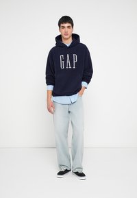 Felpa con cappuccio blu navy con logo "GAP", tessuto strutturato, tasca frontale; sovrapposta a una camicia a righe azzurro chiaro, abbinata a jeans chiari e scarpe da ginnastica nere.