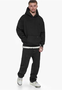 Schwarzer Kapuzenpullover mit frontaler Tasche und dezent geprägtem Logo-Design, kombiniert mit verwaschenen schwarzen Jeans und weißen Sneakers. Einfacher, lässiger Look.