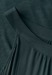 Tessuto verde scuro con una texture liscia, caratterizzato da un design drappeggiato e pieghe. Il décolleté presenta un dettaglio di spalline larghe e strutturate.
