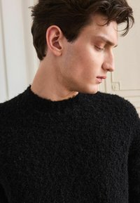 Jonge man met kort donker haar, gekleed in een gestructureerde zwarte trui, die naar beneden en opzij kijkt tegen een lichte binnenachtergrond.