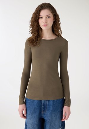 Giovane con capelli ricci che indossa una maglietta a maniche lunghe verde oliva e jeans di denim blu, in piedi davanti a uno sfondo bianco uniforme.