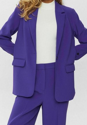 Femme portant un blazer violet et un pantalon assorti avec un haut blanc côtelé à col montant, mains sur les hanches, sur un fond uni.