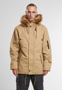 Khaki parka jakke med fôr i kunstpels, klafflommer foran, justerbar snor i hemmen og glidelåslomme på armen.