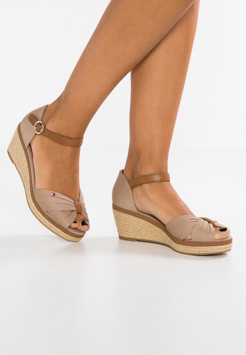 Jambes portant des espadrilles compensées beige avec brides autour de la cheville et détail noué à l'avant, sur une surface blanche réfléchissante.