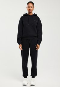 Naine seisab sirgelt, kandmas musta kapuutsiga pusa ja sobivaid dressipükse valgete tossudega heledal taustal.