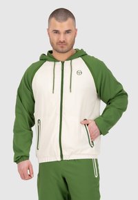 Giacca verde e crema con zip e cappuccio, maniche raglan a contrasto e due tasche con zip. Materiale sintetico con texture liscia.