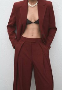 Bordeaux colbert op maat met een revers, gecombineerd met high-waisted broeken. Een zwart bikini-top en parelketting ronden de look af.