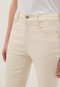 Jeans beige de cintura alta con botón y trabillas, usados por una persona con camisa blanca, con las manos relajadas a los lados.