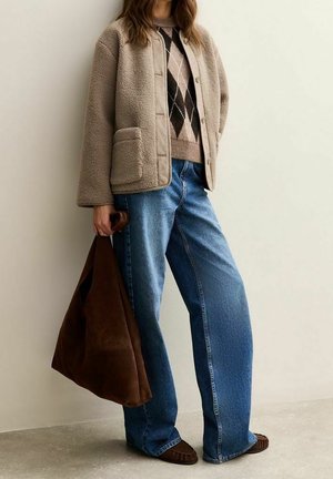 Personne portant une veste polaire beige sur un pull à losanges, jean bleu à jambes larges, mocassins marron, tenant un grand sac en daim marron.