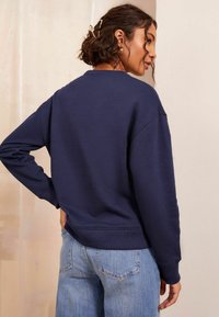 Laivansinis sweatshirt pyöreällä kaula-aukolla, ribbaantuilla hihansuilla ja helman reunalla. Kangas näyttää pehmeältä ja teksturoidulta, yhdistettynä vaaleansinisiin farkkuihin.
