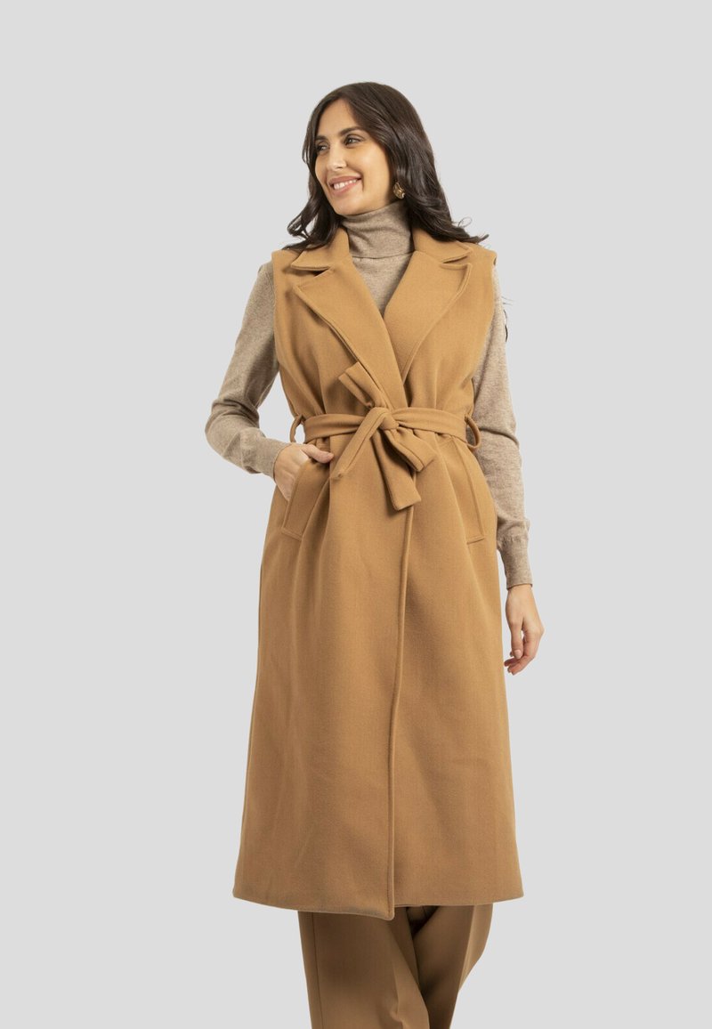 Gilet en laine sans manches couleur beige, avec une taille ceinturée, un col cranté et des poches avant. Porté sur un pull à col montant beige, texture lisse.