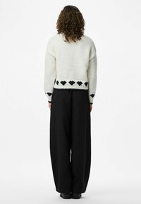 Käiv külgedega isik, kellel on lokkis juuksed, kannab valget kampsunit mustade südameprindiga ja laiu musti pükse, seistes valgel taustal seljaga.