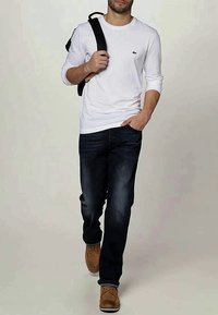 Camisa branca de manga longa, jeans azul escuro, sapatos castanhos. Mochila preta pendurada sobre o ombro esquerdo. Design simples, ajuste casual.
