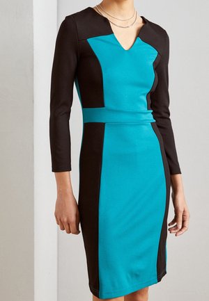 Kobieta w dopasowanej sukience do kolan z czarnymi i turkusowymi panelami w stylu color-block oraz rękawami do połowy przedramienia, ozdobiona srebrnymi łańcuszkami.