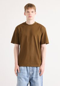 Braunes Baumwoll-T-Shirt mit kurzen Ärmeln und Rundhalsausschnitt. Mit dezent eingeprägtem Logo auf der Brust. Kombiniert mit hellblauen Jeans.