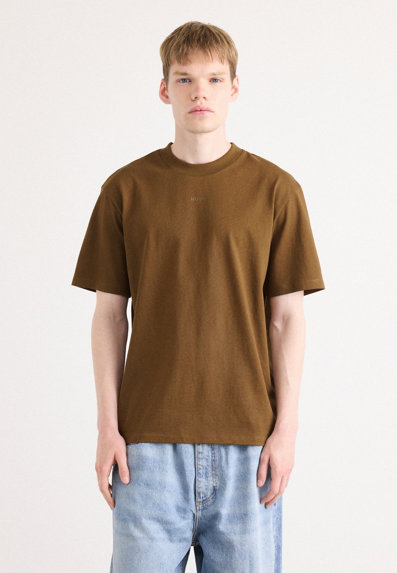Braunes Baumwoll-T-Shirt mit kurzen Ärmeln und Rundhalsausschnitt. Mit dezent eingeprägtem Logo auf der Brust. Kombiniert mit hellblauen Jeans.