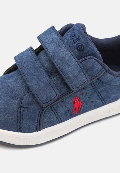 Baskets en daim bleu marine avec deux sangles Velcro ajustables, semelle en caoutchouc blanche, détails perforés et logo rouge brodé sur le côté.
