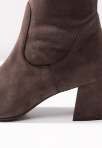 Bottine en daim marron avec un talon large et angulaire, présentant une texture lisse et un design sans couture le long du bord supérieur.