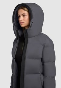Pelēks izolēts pūkains mētelis ar augstu apkakli, lielu kapuci un kontrastējošām melnām rāvējslēdzēju akcentiem; raksturīga quilted tekstūra.
