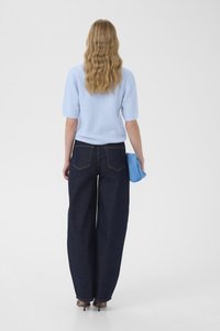 Frau mit welligem blonden Haar, die einen hellblauen Pullover, dunkle Jeans mit weitem Bein, braune High Heels trägt und eine passende blaue Clutch hält, von hinten gesehen.