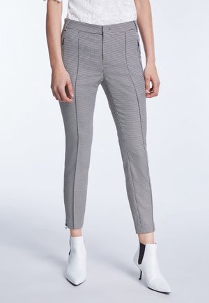 Pantalon ajusté noir et blanc à motif pied-de-poule porté avec des bottines blanches à talons et un haut en dentelle blanche visible à la taille.