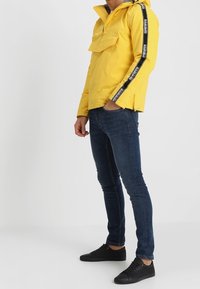 Giacca impermeabile gialla con tasca frontale, strisce laterali nere e cappuccio regolabile, abbinata a jeans blu scuro e sneakers nere.