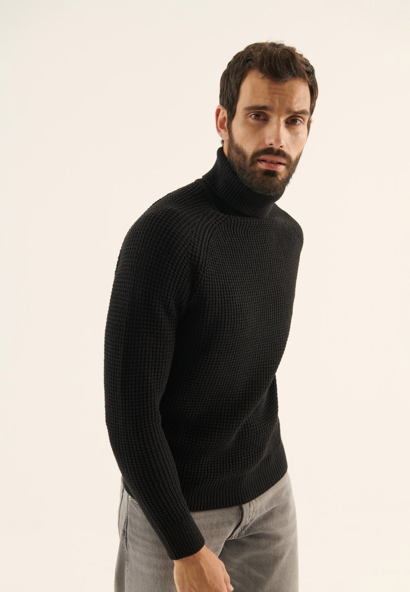 Maglione a collo alto lavorato a maglia nera con un motivo testurizzato, silhouette aderente e polsini a costine. Il modello indossa jeans grigi chiari.