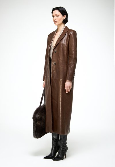 Rövid sötét hajú nő, hosszú barna bőrkabátot visel világos blúz fölött, fekete magassarkú csizmával, sötét szőrme táskát tartva a kezében.