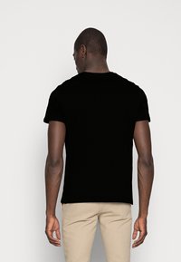Schwarzes T-Shirt mit kurzen Ärmeln und klassischem Rundhalsausschnitt, aus weichem Baumwollmaterial. Getragen mit hellfarbigen Hosen, die eine glatte Textur zeigen.